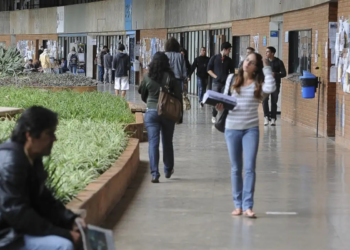 Jovens caminham e conversam em corredor amplo com jardins internos e murais repletos de avisos