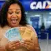 Beneficiária do Mães de Pernambuco segura cédulas de real em frente a uma agência da CAIXA, representando o pagamento de R$ 300.