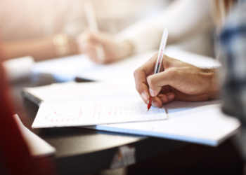 Mão masculina escreve com caneta vermelha em caderno aberto sobre mesa escura