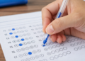 Pessoa marca respostas com caneta azul em folha de gabarito sobre mesa de madeira