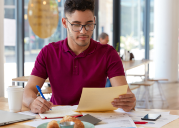 Homem com camiseta bordô anota informações em caderno enquanto segura folha amarela em mesa com café
