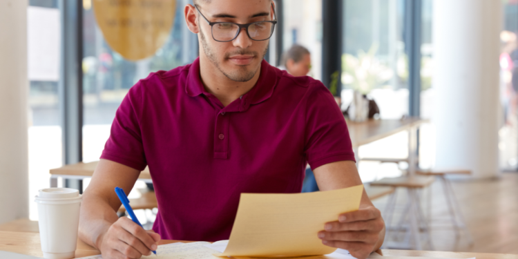 Homem com camiseta bordô anota informações em caderno enquanto segura folha amarela em mesa com café