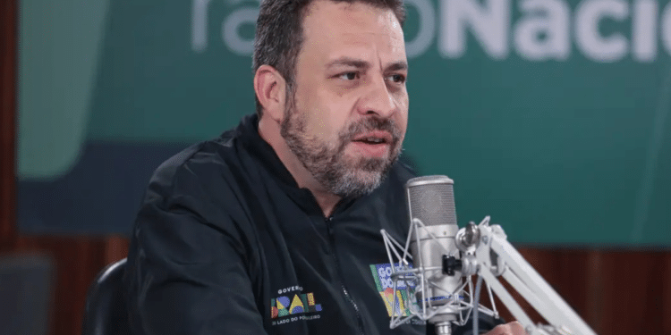 Homem de barba fala ao microfone profissional em estúdio, com jaqueta do Governo do Brasil