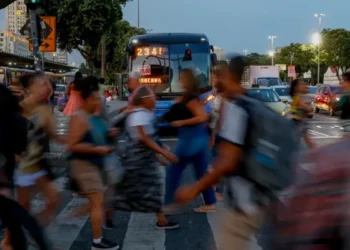 Grupo de trabalhadores atravessa faixa com ônibus e veículos ao fundo ao anoitecer