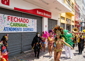 Grupo fantasiado e banda de metais desfilam em frente a agência bancária fechada durante o Carnaval