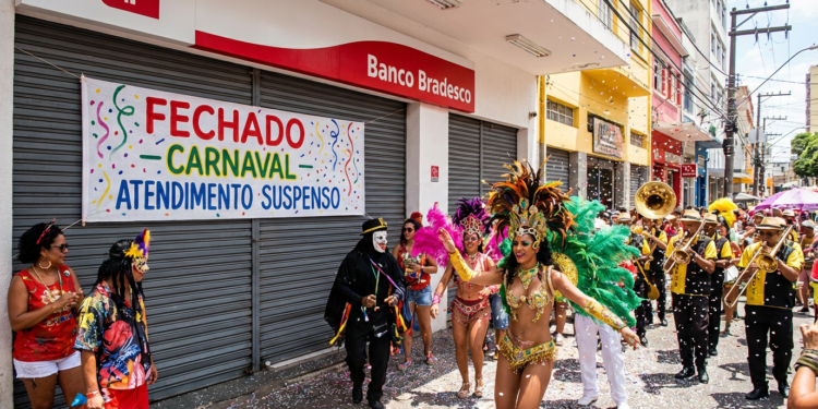 Grupo fantasiado e banda de metais desfilam em frente a agência bancária fechada durante o Carnaval