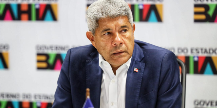 Homem de cabelos grisalhos com terno azul sentado diante de painel do Governo da Bahia