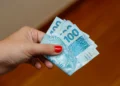 Mão segurando cédulas de cem reais representando o pagamento do programa Pé-de-Meia em 2026.