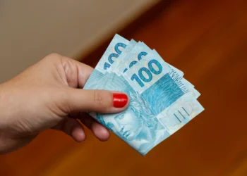 Mão segurando cédulas de cem reais representando o pagamento do programa Pé-de-Meia em 2026.