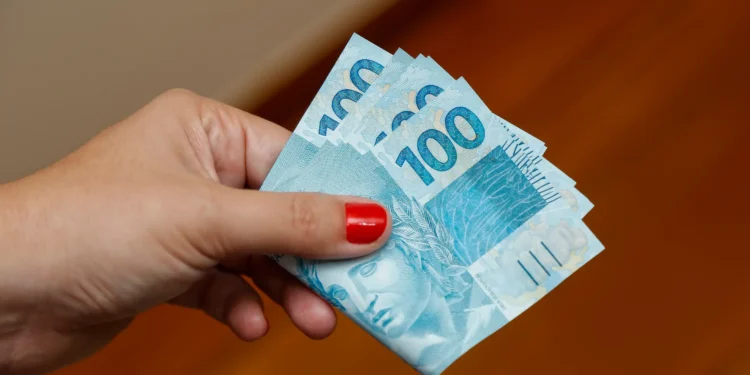 Mão segurando cédulas de cem reais representando o pagamento do programa Pé-de-Meia em 2026.