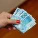 Mão segurando cédulas de cem reais representando o pagamento do programa Pé-de-Meia em 2026.
