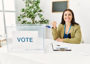 Mesária sorrindo e apontando com o polegar ao lado de uma urna eleitoral nas Eleições de 2026.