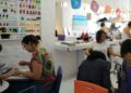 Interior de um salão de beleza movimentado com várias manicures atendendo clientes. Uma profissional à esquerda usa um smartphone enquanto trabalha, e ao fundo há uma prateleira organizada com dezenas de frascos de esmalte coloridos.
