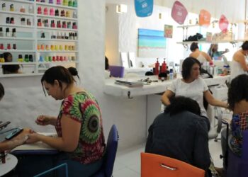 Interior de um salão de beleza movimentado com várias manicures atendendo clientes. Uma profissional à esquerda usa um smartphone enquanto trabalha, e ao fundo há uma prateleira organizada com dezenas de frascos de esmalte coloridos.