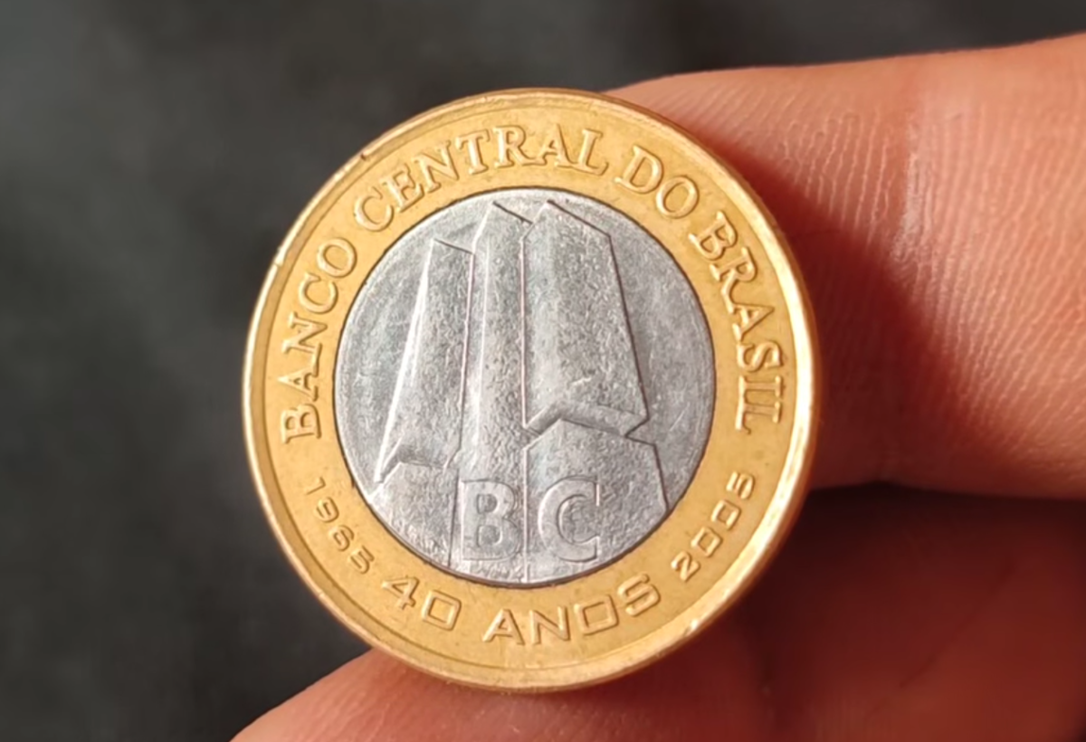 moeda 1 real bc 40 anos rara colecionismo Visão frontal detalhada da moeda de 1 real comemorativa dos 40 anos do Banco Central, segurada entre os dedos. No centro prateado, vê-se o edifício-sede do BC e a sigla "BC". No anel dourado, a inscrição "BANCO CENTRAL DO BRASIL" e "1965 40 ANOS 2005".