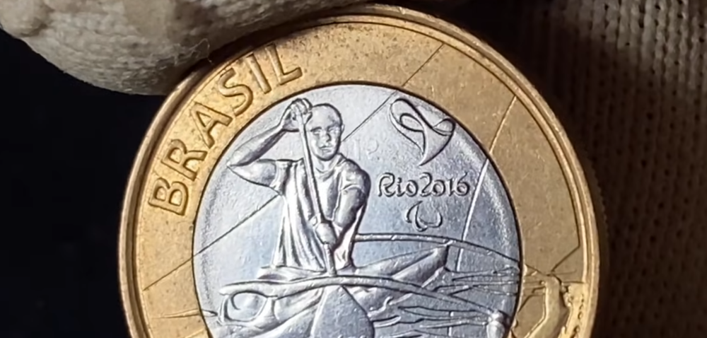 Close em alta definição de uma moeda de 1 real das Olimpíadas Rio 2016. A imagem foca no rosto do atleta e no pescoço, onde é possível notar pequenas marcas de relevo que indicam o erro de "data marcada" ou "data fantasma" em uma peça de 2015.