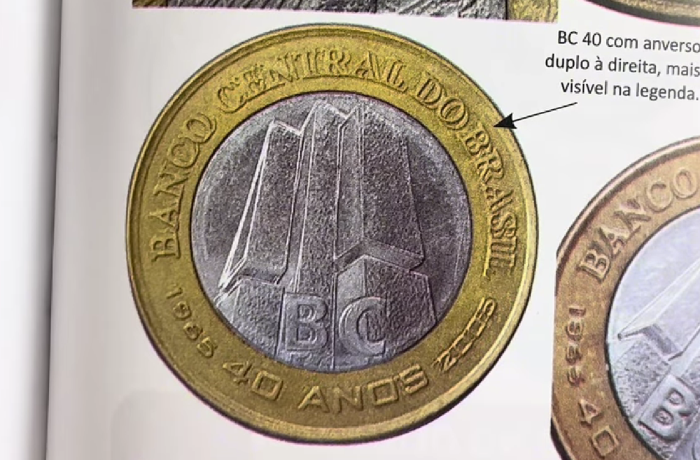 erro duplicação letras moeda 1 real bc 40 anos Close-up do anverso de uma moeda de 1 real de 2005 (BC 40 anos) apresentando erro de duplicação nas letras "BANCO CENTRAL DO BRASIL". Uma seta preta aponta para o detalhe da legenda duplicada no anel dourado à direita. A peça exibe o prédio do Banco Central no centro prateado.