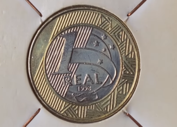 Fotografia em macro de uma moeda de 1 real brasileira do ano de 1998, centralizada em um suporte de papel branco (coin holder). A imagem destaca o número "1", a palavra "REAL" e o ano "1998" gravados no núcleo prateado, rodeado pelo anel dourado com grafismos indígenas marajoaras e estrelas do Cruzeiro do Sul.