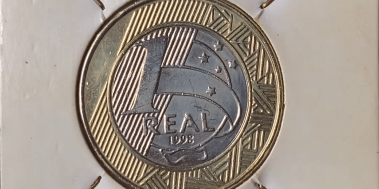 Fotografia em macro de uma moeda de 1 real brasileira do ano de 1998, centralizada em um suporte de papel branco (coin holder). A imagem destaca o número "1", a palavra "REAL" e o ano "1998" gravados no núcleo prateado, rodeado pelo anel dourado com grafismos indígenas marajoaras e estrelas do Cruzeiro do Sul.