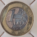 Fotografia em macro de uma moeda de 1 real brasileira do ano de 1998, centralizada em um suporte de papel branco (coin holder). A imagem destaca o número "1", a palavra "REAL" e o ano "1998" gravados no núcleo prateado, rodeado pelo anel dourado com grafismos indígenas marajoaras e estrelas do Cruzeiro do Sul.