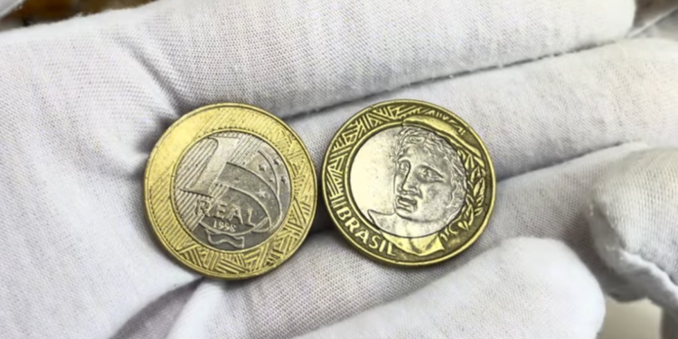 Duas mãos usando luvas brancas de tecido seguram duas moedas de 1 real de 1998. A moeda da esquerda mostra o valor "1 REAL" e o ano "1998" no centro de cupro-níquel, cercado por um anel de alpaca dourado. A moeda da direita mostra a efígie da República voltada para a esquerda.