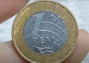 Close-up de uma moeda de 1 Real do ano de 2008, segurada entre os dedos, mostrando a face do valor com a data para identificação da raridade.