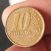 Close de uma moeda de 10 centavos de real do ano de 2002, fabricada em aço revestido de bronze, sendo segurada pelos dedos de uma pessoa.