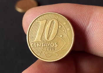 Close-up frontal de uma moeda dourada de 10 centavos de Real sendo segurada por dedos, com foco nítido no ano de fabricação 1999 e no numeral 10.