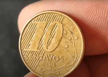 Foto em detalhe do reverso de uma moeda de 10 centavos de real do ano de 2003, segurada por dedos. A imagem mostra o numeral "10", a palavra "CENTAVOS" e o ano "2003" sobre um fundo de linhas diagonais e constelação do Cruzeiro do Sul.