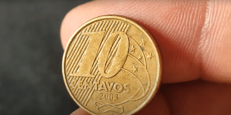Foto em detalhe do reverso de uma moeda de 10 centavos de real do ano de 2003, segurada por dedos. A imagem mostra o numeral "10", a palavra "CENTAVOS" e o ano "2003" sobre um fundo de linhas diagonais e constelação do Cruzeiro do Sul.