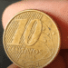 Foto em detalhe do reverso de uma moeda de 10 centavos de real do ano de 2003, segurada por dedos. A imagem mostra o numeral "10", a palavra "CENTAVOS" e o ano "2003" sobre um fundo de linhas diagonais e constelação do Cruzeiro do Sul.