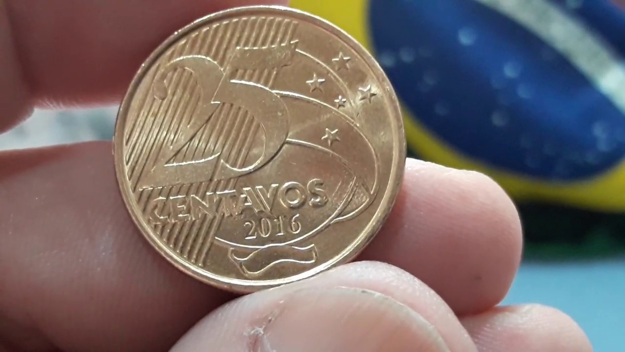 moeda 25 centavos 2016 baixa tiragem valor Close-up da mão de uma pessoa segurando uma moeda de 25 centavos de 2016. A moeda é dourada, está bem conservada e o ano de cunhagem 2016 aparece nítido na parte inferior. Ao fundo, as cores verde e amarelo da bandeira do Brasil estão desfocadas.
