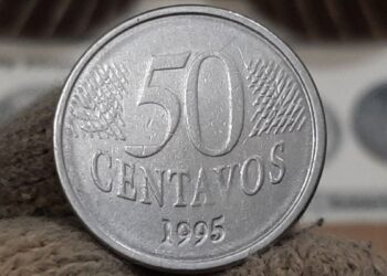 Mão com luva de tecido branca segurando uma moeda de 50 centavos de 1995 entre o polegar e o indicador. Ao fundo, outros exemplares de moedas raras aparecem desfocados em suportes de proteção prateados, destacando o foco na peça central.