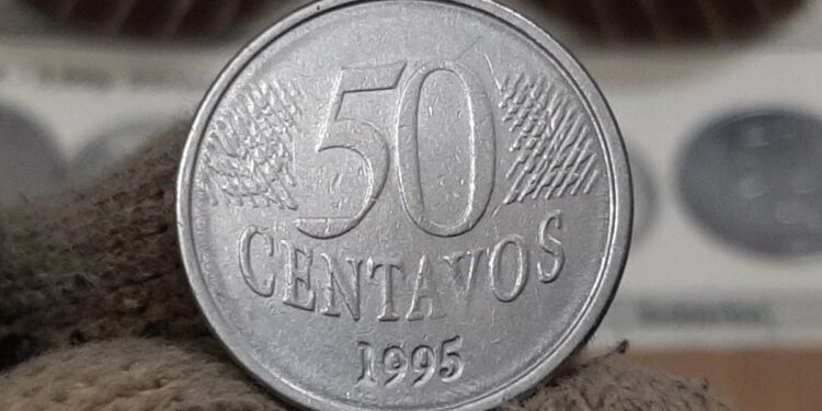 Mão com luva de tecido branca segurando uma moeda de 50 centavos de 1995 entre o polegar e o indicador. Ao fundo, outros exemplares de moedas raras aparecem desfocados em suportes de proteção prateados, destacando o foco na peça central.