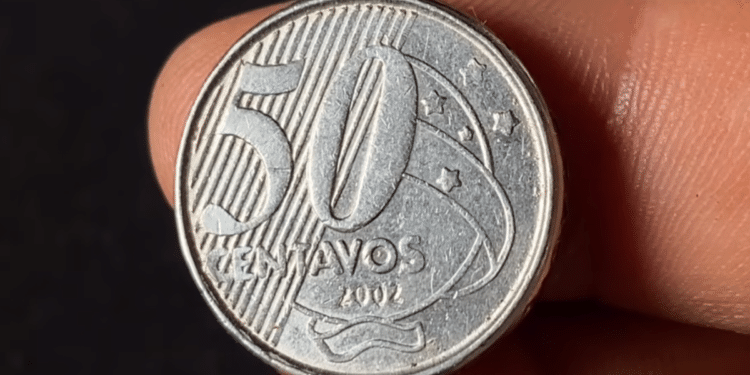 Close-up detalhado de uma moeda de 50 centavos de real do ano 2002, fabricada em aço inoxidável, sendo segurada entre os dedos contra um fundo escuro.
