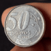 Close-up detalhado de uma moeda de 50 centavos de real do ano 2002, fabricada em aço inoxidável, sendo segurada entre os dedos contra um fundo escuro.