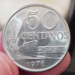 Close-up do reverso de uma moeda de 50 centavos de 1976, segurada por dedos em primeiro plano. O design apresenta o valor "50 CENTAVOS", o ano "1976" na base e a imagem de um navio cargueiro navegando, com detalhes nítidos do metal prateado.