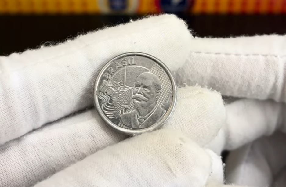 moeda 50 centavos 2020 defeito cavanhaque valor Close-up de uma mão usando luva de tecido branco segurando uma moeda de 50 centavos do Real. O detalhe foca no rosto do Barão do Rio Branco, onde é possível ver uma pequena marca de metal elevada no queixo, caracterizando o erro de cunho quebrado conhecido como "cavanhaque".