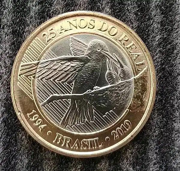 moeda 1 real 25 anos plano real detalhes Close-up de uma moeda de 1 real de 2019, comemorativa de 25 anos do Plano Real, com brilho metálico e detalhes nítidos do beija-flor no ninho.