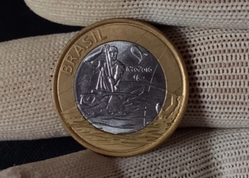 Close do anverso de uma moeda de 1 real das Olimpíadas Rio 2016, modalidade Paracanoagem. A imagem mostra o atleta remando em destaque prateado sobre o núcleo da moeda, segurada por dedos protegidos com luvas de tecido branco.