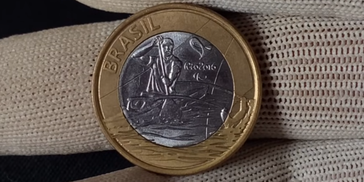 Close do anverso de uma moeda de 1 real das Olimpíadas Rio 2016, modalidade Paracanoagem. A imagem mostra o atleta remando em destaque prateado sobre o núcleo da moeda, segurada por dedos protegidos com luvas de tecido branco.