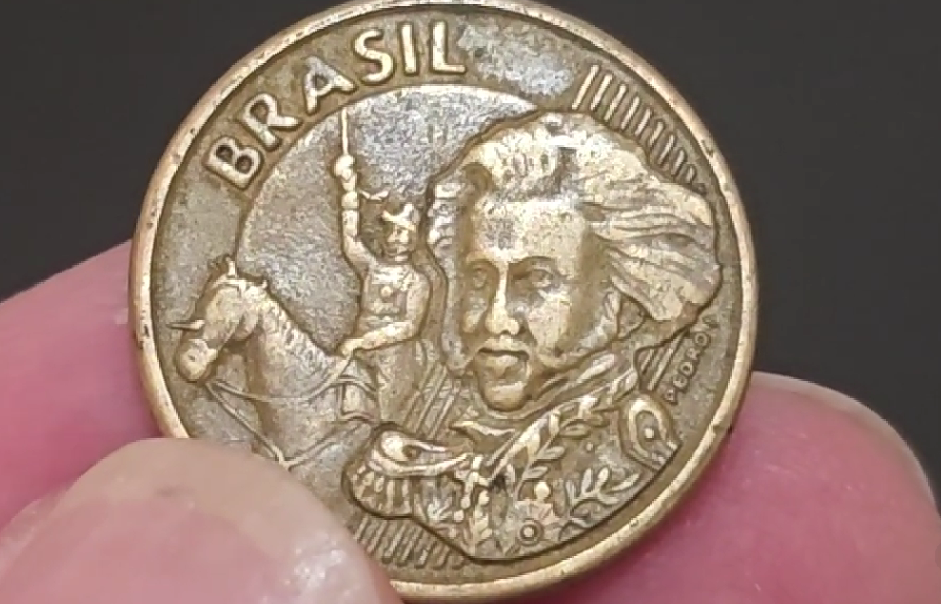 identificar moeda 10 centavos 2003 ppp unicórnio Close-up de uma moeda de 10 centavos de 2003 sendo segurada por dedos. A imagem destaca a efígie de Dom Pedro I e um cavalo ao fundo. É possível observar o detalhe do erro "PPP" (Ponto no Peito) e a variante "Unicórnio" com uma saliência na cabeça do animal.