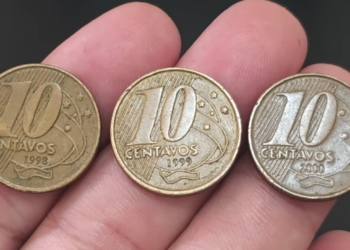 Close de uma mão segurando três moedas de 10 centavos da segunda família do Real, alinhadas lado a lado. As moedas exibem claramente os anos de cunhagem 1998, 1999 e 2000, da esquerda para a direita.