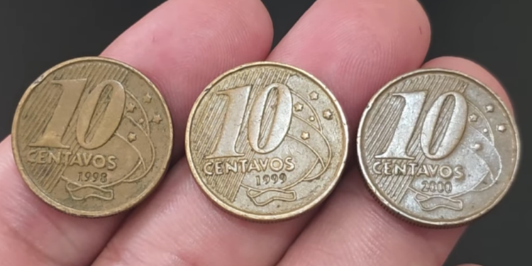 Close de uma mão segurando três moedas de 10 centavos da segunda família do Real, alinhadas lado a lado. As moedas exibem claramente os anos de cunhagem 1998, 1999 e 2000, da esquerda para a direita.