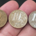 Close de uma mão segurando três moedas de 10 centavos da segunda família do Real, alinhadas lado a lado. As moedas exibem claramente os anos de cunhagem 1998, 1999 e 2000, da esquerda para a direita.