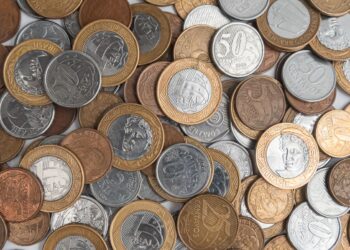 Diversas moedas do Plano Real espalhadas e sobrepostas, incluindo moedas de 1 Real, 50 centavos e 10 centavos de diferentes anos e metais.