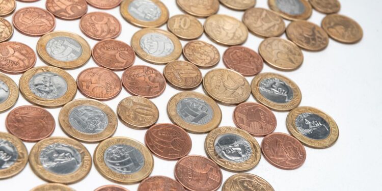 Vista superior de dezenas de moedas brasileiras de diversos valores, como 5, 10, 50 centavos e 1 Real, espalhadas sobre uma superfície branca lisa. As moedas apresentam diferentes tonalidades de dourado, prateado e cobre.
