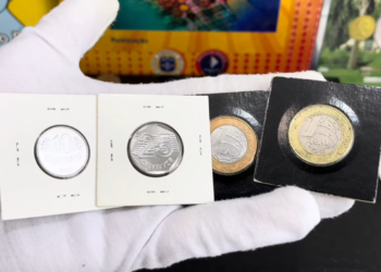 Mão usando luva branca de colecionador segurando quatro moedas raras do Real protegidas por suportes de papelão (coin holders). Estão visíveis as moedas de 10 centavos, 25 centavos e duas de 1 Real.