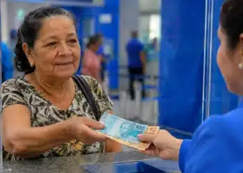 Aposentada recebe cédulas de real durante atendimento em uma agência bancária, representando os pagamentos de até R$ 8.475,55 previstos para julho.