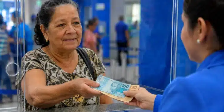 Aposentada recebe cédulas de real durante atendimento em uma agência bancária, representando os pagamentos de até R$ 8.475,55 previstos para julho.